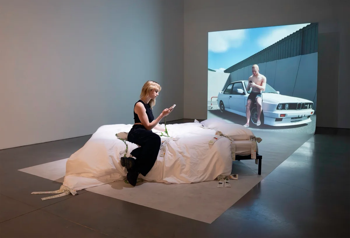 The artist sits on a bed draped in white sheets